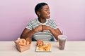 Young african american woman eating a tasty classic burger with fries and soda looking away to side with smile on face, natural Royalty Free Stock Photo