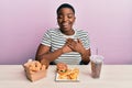Young african american woman eating a tasty classic burger with fries and soda smiling with hands on chest with closed eyes and