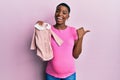 Young african american woman expecting a baby holding clothes pointing thumb up to the side smiling happy with open mouth Royalty Free Stock Photo