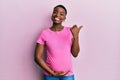 Young african american woman expecting a baby, touching pregnant belly pointing to the back behind with hand and thumbs up, Royalty Free Stock Photo