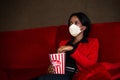 Young African American woman in formal business suit wearing a protective mask in movie theater. She& x27;s eating a popcorn Royalty Free Stock Photo