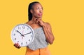 Young african american woman holding big clock serious face thinking about question with hand on chin, thoughtful about confusing Royalty Free Stock Photo