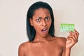Young african american woman holding birth control pills in shock face, looking skeptical and sarcastic, surprised with open mouth Royalty Free Stock Photo