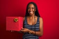 Young african american woman holding birthday gift standing over isolated red background with a happy face standing and smiling Royalty Free Stock Photo