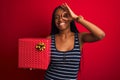 Young african american woman holding birthday gift standing over isolated red background with happy face smiling doing ok sign Royalty Free Stock Photo