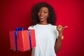 Young african american woman holding birthday gift standing over isolated red background pointing and showing with thumb up to the Royalty Free Stock Photo