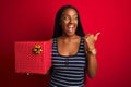 Young african american woman holding birthday gift standing over isolated red background pointing and showing with thumb up to the Royalty Free Stock Photo