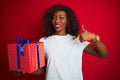 Young african american woman holding birthday gift standing over isolated red background with surprise face pointing finger to Royalty Free Stock Photo