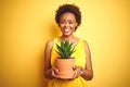 Young african american woman holding cactus pot over isolated yellow background with a happy face standing and smiling with a Royalty Free Stock Photo