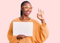 Young african american woman holding cardboard banner with blank space doing ok sign with fingers, smiling friendly gesturing Royalty Free Stock Photo