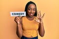 Young african american woman holding comedy word paper doing ok sign with fingers, smiling friendly gesturing excellent symbol