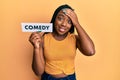 Young african american woman holding comedy word paper stressed and frustrated with hand on head, surprised and angry face Royalty Free Stock Photo