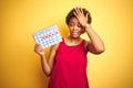Young african american woman holding menstruation calendar over isolated yellow background stressed with hand on head, shocked