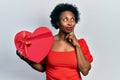 Young african american woman holding valentine gift serious face thinking about question with hand on chin, thoughtful about