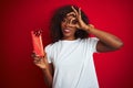 Young african american woman holding valentine gift standing over isolated red background with happy face smiling doing ok sign Royalty Free Stock Photo