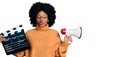 Young african american woman holding video film clapboard and megaphone making fish face with mouth and squinting eyes, crazy and Royalty Free Stock Photo