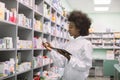 Young African American woman pharmacist or chemist using digital tablet, while standing near the shelves with medicines Royalty Free Stock Photo