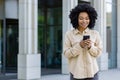 Young African American woman with phone smiling, businesswoman outside office building uses smartphone app, types Royalty Free Stock Photo