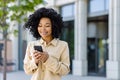 Young African American woman with phone smiling, businesswoman outside office building uses smartphone app, types Royalty Free Stock Photo