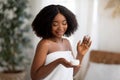 Young African American woman posing with jar of hydrating cream, smiling, wearing bath towel indoors Royalty Free Stock Photo
