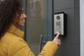 Young African-American woman ringing intercom with camera near building entrance