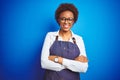 Young african american woman shop owner wearing business apron over blue background happy face smiling with crossed arms looking Royalty Free Stock Photo