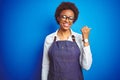 Young african american woman shop owner wearing business apron over blue background smiling with happy face looking and pointing Royalty Free Stock Photo