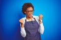 Young african american woman shop owner wearing business apron over blue background very happy and excited doing winner gesture