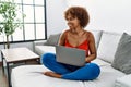 Young african american woman sitting on the sofa at home using laptop looking away to side with smile on face, natural expression Royalty Free Stock Photo