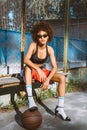 Young african-american woman in sportive attire and high heels sitting on bench with basketball ball under Royalty Free Stock Photo