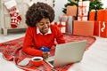 Young african american woman using laptop and credit card lying by christmas tree at home Royalty Free Stock Photo