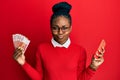 Young african american woman using smartphone holding norwegian krone banknotes puffing cheeks with funny face Royalty Free Stock Photo