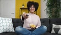 Young african american woman watching tv sitting on sofa eating chips at home Royalty Free Stock Photo