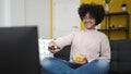 Young african american woman watching tv sitting on sofa eating chips at home Royalty Free Stock Photo