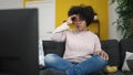 Young african american woman watching tv sitting on sofa eating chips at home Royalty Free Stock Photo