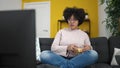 Young african american woman watching tv sitting on sofa eating chips at home Royalty Free Stock Photo