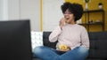 Young african american woman watching tv sitting on sofa eating chips at home Royalty Free Stock Photo