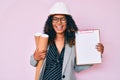 Young african american woman wearing architect hardhat holding blueprints and clipboard winking looking at the camera with sexy Royalty Free Stock Photo