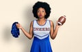 Young african american woman wearing cheerleader uniform holding pompom and football ball puffing cheeks with funny face