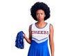 Young african american woman wearing cheerleader uniform holding pompom puffing cheeks with funny face