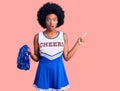 Young african american woman wearing cheerleader uniform holding pompom surprised pointing with finger to the side, open mouth Royalty Free Stock Photo