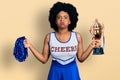 Young african american woman wearing cheerleader uniform holding pompom and trophy puffing cheeks with funny face