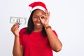 Young african american woman wearing christmas hat and holding one dollar banknote with happy face smiling doing ok sign with hand Royalty Free Stock Photo