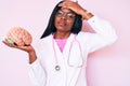 Young african american woman wearing doctor coat holding brain stressed and frustrated with hand on head, surprised and angry face Royalty Free Stock Photo