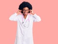 Young african american woman wearing doctor coat and stethoscope smiling pulling ears with fingers, funny gesture