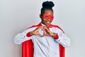 Young african american woman wearing doctor uniform and super hero costume smiling in love doing heart symbol shape with hands Royalty Free Stock Photo