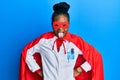 Young african american woman wearing doctor uniform and super hero costume sticking tongue out happy with funny expression