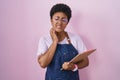 Young african american woman wearing professional waitress apron holding clipboard touching mouth with hand with painful