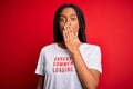 Young african american woman wearing sarcasm coments text on t-shirt over red background cover mouth with hand shocked with shame