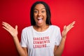 Young african american woman wearing sarcasm coments text on t-shirt over red background very happy and excited, winner expression